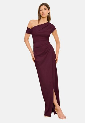 Kvinde iført en ærmeløs, skulderfri, mørkelilla aftenkjole med højt slid, stående mod en ensfarvet hvid baggrund.