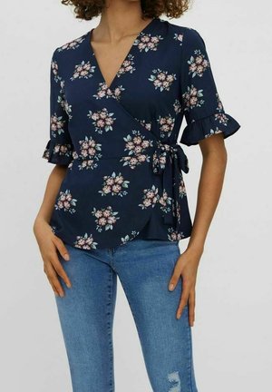 Woman wearing a navy floral wrap blouse with ruffled sleeves and light blue distressed jeans, standing with hands relaxed by sides.