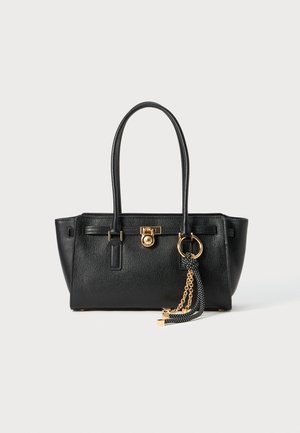 Black leather handbag with double handles, gold lock and chain detail, and black-and-white tassel accessory against white background.