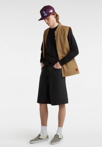 Chaleco sin mangas color bronce sobre una camisa negra de manga larga, combinado con pantalones cortos de mezclilla negros y zapatillas de cuadros. Gorra morada con gráficos.