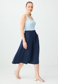 Femme marchant, portant un haut sans manches bleu clair, une jupe midi bleu marine avec poches, et des sandales blanches à talons, sur un fond clair uni.