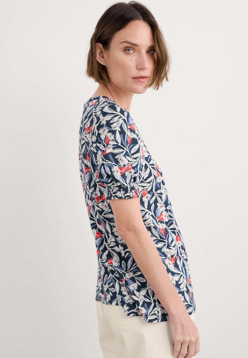 Ziedu raksta tops ar īsām pūkām, ar tumši zilu fona krāsu, sarkaniem un baltajiem ziediem, brīvu piegriezumu un sānu šķēlumiem, lai nodrošinātu vieglu kustību.