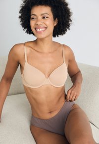 Beige onderwired bra met gladde cups en verstelbare bandjes, gecombineerd met bruine high-waisted slips. Model zit op een lichtgekleurde bank.