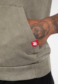 Olijfgroene hoodie met een voorzak, ribbelboorden en een klein rood merkteken. Zacht katoenmateriaal met een licht versleten textuur.