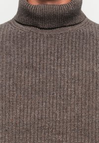 Närbild av en person som bär en tjock, ribbad brun polotröja med synlig stickad textur och vikta krage.