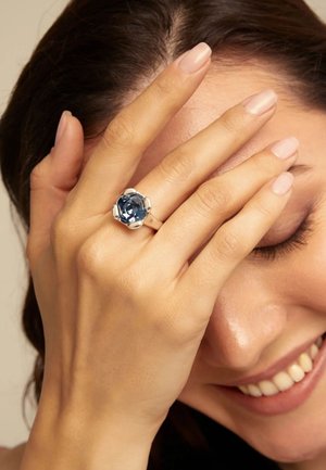 Mano con uñas de rosa pálido que lleva un anillo de plata con una gran piedra preciosa azul oscuro sobre el rostro sonriente de una mujer, ojos cerrados.