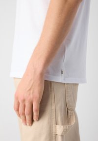 Hvid bomulds t-shirt med rund hals og korte ærmer, med et lille mærke på siden. Beige bukser med sidelommer.