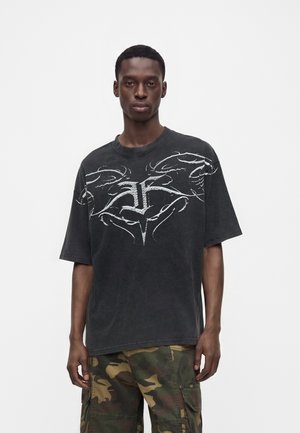 Ung mand iført en sort grafisk t-shirt og camouflage cargobukser stående mod en ensfarvet hvid baggrund.