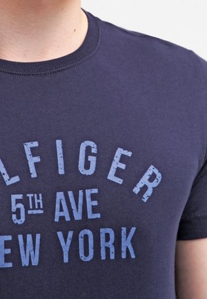 Personne portant un t-shirt bleu foncé avec un texte bleu clair usé indiquant "TOMMY HILFIGER 5th AVE NEW YORK" sur la poitrine.