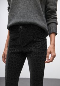 Pantaloni con stampa leopardata nera realizzati in tessuto morbido, dotati di tasche frontali e chiusura con bottone. Abbinati a un maglione grigio con polsini a coste.