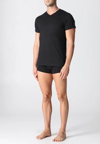 T-shirt nera a maniche corte con scollo a V realizzata in tessuto elasticizzato; design aderente. Modello abbinato a biancheria intima nera e scalzo, in piedi contro uno sfondo neutro.