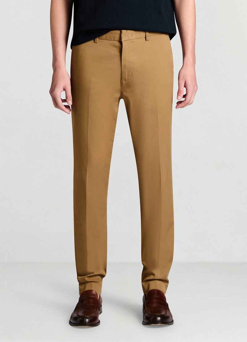 Maßgeschneiderte Hose aus einem hellbraunen Stoff. Sie zeichnet sich durch eine glatte Textur, einen geraden Schnitt und eine saubere Vorderseite ohne sichtbare Taschen aus. Kombiniert mit dunklen Schuhen.