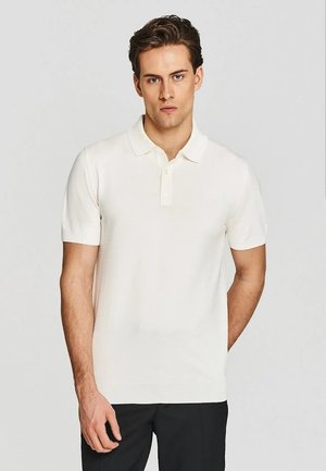 Młody mężczyzna o krótkich brązowych włosach, ubrany w białą koszulkę polo z krótkim rękawem i czarne spodnie, stoi na jednolitym jasnym tle.