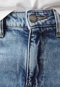 Gros plan sur un jean en denim bleu avec des coutures marron et un bouton en métal embossé du nom de la marque Lee.