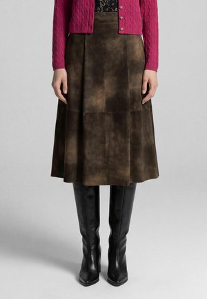 Frau trägt einen dunkelbraunen Wildleder-Midirock, schwarze kniehohe Lederstiefel und eine rote gestrickte, geknöpfte Strickjacke.