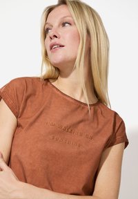 Braunes T-Shirt mit kurzen Ärmeln und rundem Ausschnitt, auf dem der Text "I'M WALKING ON SUNSHINE" in einem helleren Braun gestickt ist. Weicher Stoff.
