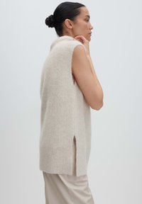 Femme aux cheveux bruns relevés en chignon, portant un gilet sans manches en tricot beige et un pantalon de couleur claire, se tenant de profil contre un fond uni.