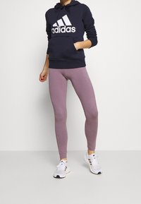 Granatowa bluza z kapturem z białym logo "adidas", z przednią kieszenią, w połączeniu z jasnofioletowymi legginsami i białymi butami sportowymi z czarnymi akcentami.