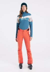 Synthetisch lange mouw top in blauw met colorblock ontwerp; oranje skibroek met zwarte accenten; zwarte skischoenen met gespen.