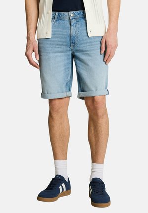 Mann trägt hellblaue, hochgekrempelte Jeansshorts, weiße Socken und marineblaue Sneakers mit weißen Streifen, steht mit entspannt an den Seiten hängenden Händen.