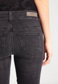 Jeans de color gris oscuro con un diseño ajustado, que cuentan con un diseño de cinco bolsillos, costuras definidas y una etiqueta de cuero de marca en la cinturilla.