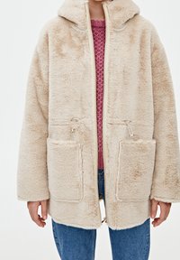 Beige fuzzy jakke med hætte, lynlås foran og to forlommer. Tykt materiale med en afslappet pasform, bæres over en lyserød sweater og jeans.