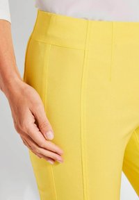 Gele stoffen broek met een hoge taille, slanke silhouet en naden aan de voorkant; hand op de heup toont textuur en pasvorm.
