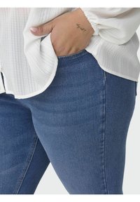 Bijela teksturirana bluzica s naglašenim rukavima u kombinaciji s tamnoplavim uskim trapericama, koje prikazuju glatki denim i suptilne detalje šivanja.