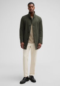 Donkergroene jas met hoge kraag, knoopsluiting en zijzakken, gecombineerd met een beige top en crèmekleurige broek, aangevuld met zwarte schoenen.