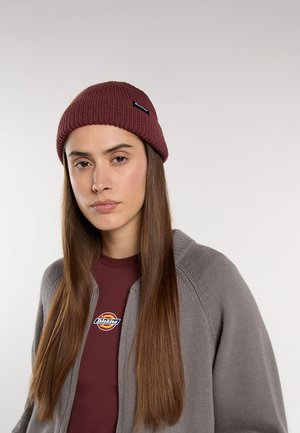 Berretto lavorato a maglia borgogna con texture a coste e un'etichetta nera. Indossato con una camicia bordeaux e un cardigan grigio, caratterizzato da un logo rotondo. Capelli lunghi in vista.