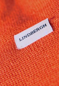 Jasnopomarańczowa dziana tkanina o drobnej fakturze i widocznym szwie. Biała metka z napisem "LINDBERGH" w czarnym kolorze.