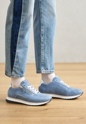 Person trägt hellblaue Sneakers mit weißen Schnürsenkeln und Reißverschlüssen, kombiniert mit hellblauen Jeans, steht auf einem Holzboden.
