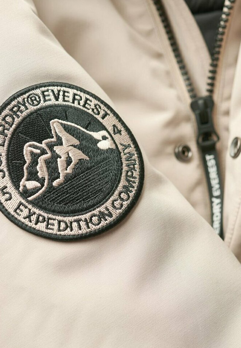 Beige Jackenstoff mit einem schwarzen runden gestickten Aufnäher, der Berggrafiken und den Text "EVERST EXPEDITION COMPANY" zeigt. Reißverschlussdetails sind sichtbar.