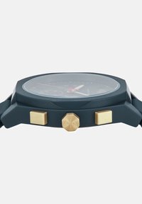 Muški sat s okruglim, crnim plastičnim kućištem, zlatnim akcentima i crnim brojčanikom. Ima naglašene kazaljke i sub-brojčanike za kronograf. Gumeni remen.