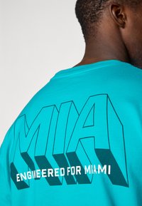 Tyrkysové bavlněné tričko s výrazným černým obrysem grafiky "MIA" a textem "ENGINEERED FOR MIAMI". Krátké rukávy a kulatý výstřih.