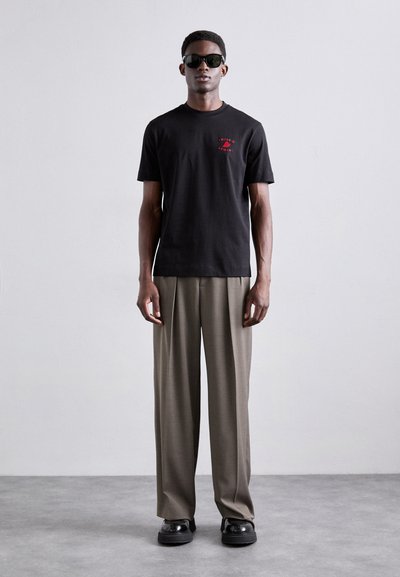 T-shirt en coton noir avec logo rouge, associé à un pantalon beige large et des chaussures noires brillantes. Le mannequin porte des lunettes de soleil noires.