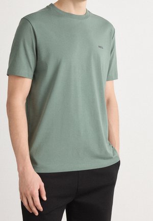 Muž v světle zeleném tričku s krátkými rukávy a logem "BOSS" na hrudi, v černých kalhotách, stojící před jednoduchým pozadím.