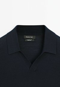 Námořnický modrý merinový svetr s výstřihem do V a límcem. Má hladkou texturu a detaily štítku na výstřihu. Jednoduchý a klasický design.