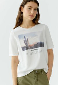 Weißes Baumwoll-T-Shirt mit einem Kaktus-Sonnenuntergangs-Grafik und Text, Rundhalsausschnitt und kurzen Ärmeln, kombiniert mit olivgrünen Hosen.