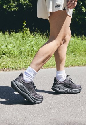 Legs of a person walking outdoors wearing black HOKA sneakers, white socks, and a white skirt on a paved path with grass background.