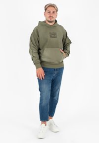 Olijfgroene hoodie met een voorkantzak en reliëflogo, gecombineerd met lichtblauwe jeans en witte sneakers, tegen een effen achtergrond.