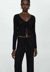 Čierny pletený crop top a zodpovedajúce nohavice s detailom na viazanie vpredu, dlhými rukávmi a vertikálnymi pruhmi, predvádzajúci ľahkú, textúrovanú tkaninu.