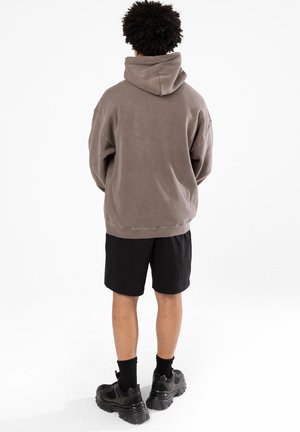 Przesadnie duża brązowa bluza polarowa z wysokim kołnierzem, zestawiona z czarnymi szortami i czarnymi trampkami. Materiał wydaje się miękki i o fakturze.