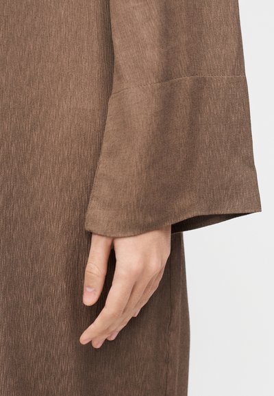 Χέρι που ξεκουράζεται δίπλα σε καφετί πολύχρωμο ρούχο με μακριά μανίκια και φαρδύ, ελαφρώς φουσκωτό μανσέτα, πάνω σε μονόχρωμο φόντο.