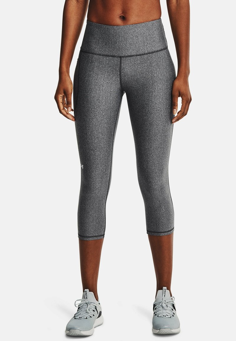 Personne portant des leggings gris taille moyenne coupés et des chaussures de sport gris clair, debout devant un fond uni.