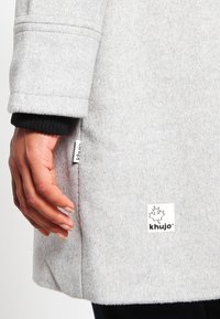 Cappotto di lana grigio con polsini neri a coste. Presenta un'etichetta laterale e un materiale testurizzato. Mano appoggiata sul cappotto.