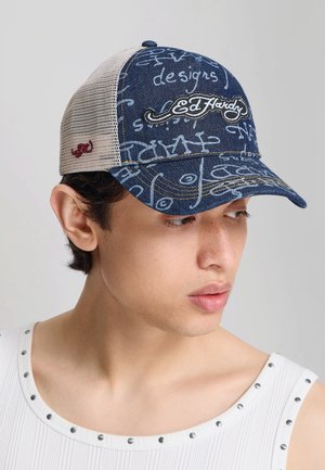 Młody mężczyzna w białym tank topie, noszący niebiesko-beżową czapkę Ed Hardy z wzorami w stylu graffiti i siateczkowym tyłem.