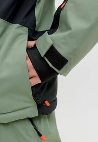 Green and black jacket featuring a smooth texture, adjustable Velcro cuffs, and orange zipper pulls. Side pocket accessed from the front.