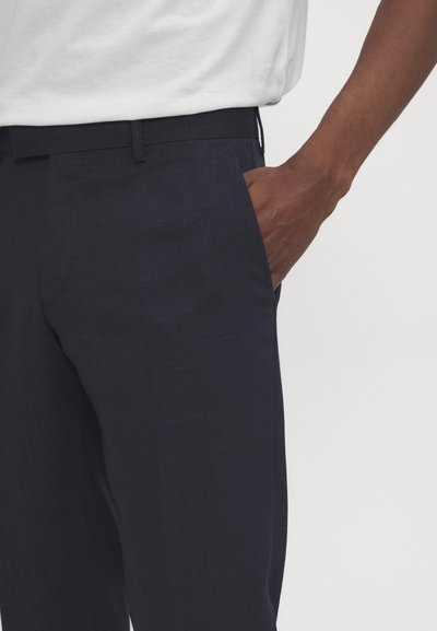Mørkeblå, slim-fit bukser laget av teksturert stoff, med en sidelomme. Brukt sammen med en hvit t-skjorte, som viser en klassisk, uformell stil.