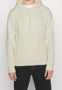 Kapturowa bluza w jasnym kremowym kolorze, z przednią kieszenią typu kangur i wytłoczonym logo "HACKETT LONDON". Miękka tkanina, swobodny krój.